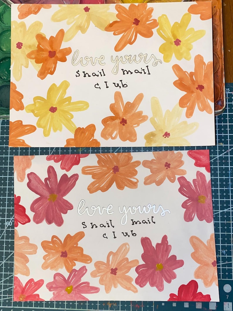 May include: Two rectangular cards featuring hand-painted floral patterns. The top card has yellow and orange flowers, while the bottom card has pink and orange flowers. Both cards display the text "love yours snail mail club" in the center.