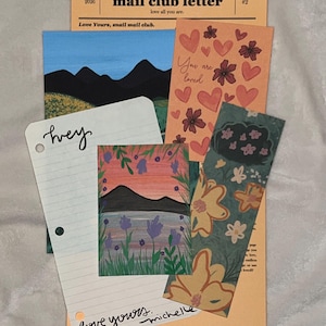May include: A collection of stationery items including a peach-colored Love Yours Snail Mail Club letter, a landscape print with mountains, a card with hearts and the text You are loved, a lined paper with the word hey and love yours michelle, and a floral bookmark.