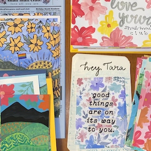 May include: A collection of stationery items including a Snail Mail Club newsletter, floral patterned cards, and a note with the text "hey, Tara" and "good things are on its way to you." The items feature colorful illustrations of flowers, landscapes, and mountains.