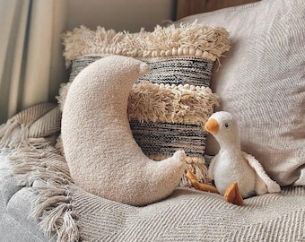 May include: A plush, cream-colored crescent moon pillow rests on a gray herringbone blanket, next to a decorative pillow with black and cream stripes and fringe. A white stuffed goose with an orange beak and feet is also present.