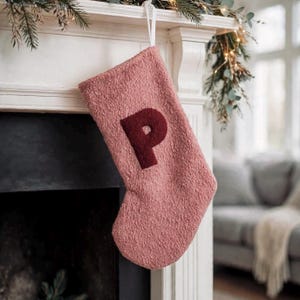 May include: A dusty rose-coloured Christmas stocking made of a textured, fuzzy material. The stocking features a large, dark burgundy letter P appliqued in the centre and includes a white fabric loop at the top for hanging purposes.