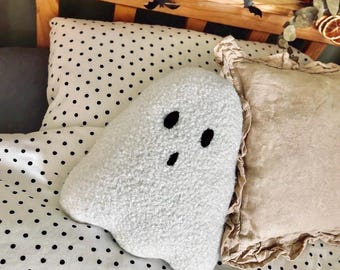 May include: A white ghost-shaped throw pillow made of textured, fuzzy sherpa fabric. The pillow features two oval-shaped black eyes and a small, vertically oriented oval-shaped black mouth embroidered onto the center of the front surface.