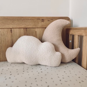 May include: Two plush pillows in the shape of a cloud and a crescent moon. The pillows are beige and white and are sitting on a white and gray patterned crib sheet.