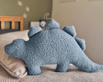 May include: A blue dinosaur-shaped throw pillow made of a textured, nubby fabric. The pillow features a long tail, four short legs, and a series of triangular plates along its back, mimicking the appearance of a stegosaurus silhouette.