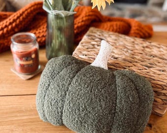 May include: A plush, olive green pumpkin-shaped decorative pillow featuring a textured, sherpa-like fabric finish and a short, cream-colored stem made of a different, lighter-toned material. The pumpkin has a rounded, segmented design.
