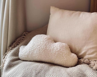 May include: A white cloud-shaped decorative pillow with a fluffy, textured surface rests on a patterned blanket. A solid, light-colored pillow is behind it. The scene evokes a cozy, comfortable atmosphere, perfect for a bedroom or living room.