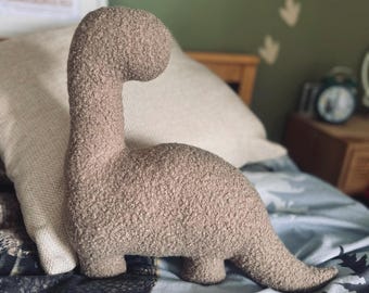 May include: A plush dinosaur toy with a textured, curly, light brown fabric surface. The dinosaur has a long, curved neck, a rounded head, a stout body, and a tapered tail, standing in a profile position with four short, thick legs.