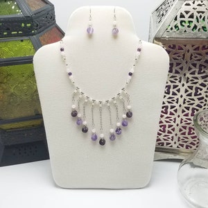 Op de afbeelding: Een zilveren ketting met een witte en paarse edelsteenfranje. De ketting wordt getoond op een witte mannequinbuste. Passende oorbellen met paarse edelstenen zijn ook inbegrepen.