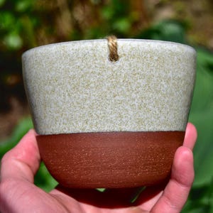 May include: A white and brown ceramic hanging planter with a rope loop. The planter has a speckled white glaze on the top half and a solid brown glaze on the bottom half.
