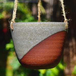 May include: A hanging planter made of ceramic with a white and brown glaze. The planter has a rope hanging system.