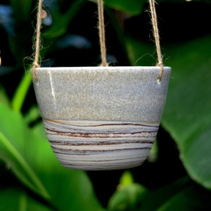 May include: A hanging planter made of ceramic with a light grey speckled glaze and brown and white stripes. The planter is suspended by twine.