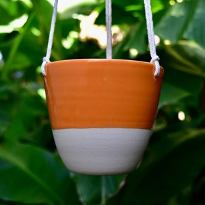 Pode incluir: Um vaso pendurado de cerâmica laranja e branca com uma corda branca.