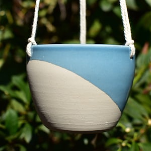 May include: A blue and white ceramic hanging planter with a rope hanger. The planter is half blue and half white, with a curved line separating the two colors.
