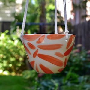 May include: A white ceramic hanging planter with a bright orange abstract floral design. The planter is suspended by white cords.