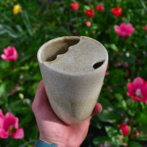 Puede incluir: Una taza de cerámica blanca con un esmalte moteado y un diseño asimétrico único. La taza tiene dos aberturas en la parte superior, creando una forma distintiva.