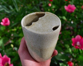 May include: A white ceramic mug with a speckled glaze and a unique, asymmetrical design. The mug has two openings on the top, creating a distinctive shape.