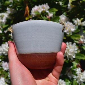 Puede incluir: Un peque&ntilde;o macetero de cer&aacute;mica gris claro con un borde marr&oacute;n y un asa de cuerda natural. El macetero se est&aacute; sosteniendo en una mano contra un fondo de flores blancas.