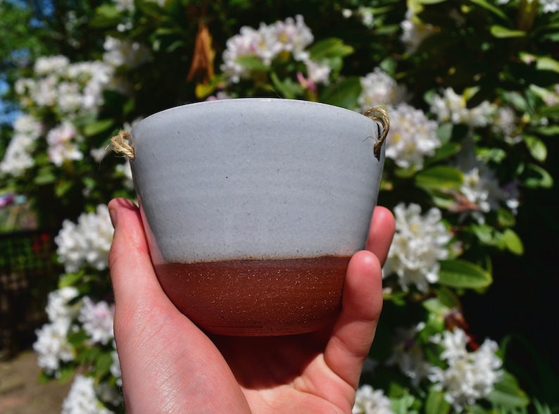 Puede incluir: Un peque&ntilde;o macetero de cer&aacute;mica gris claro con un borde marr&oacute;n y un asa de cuerda natural. El macetero se est&aacute; sosteniendo en una mano contra un fondo de flores blancas.
