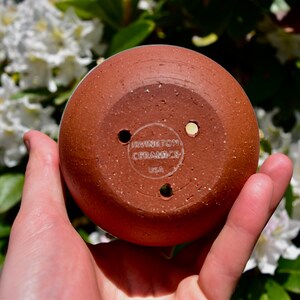 Puede incluir: Una maceta de cer&aacute;mica de color terracota con tres agujeros de drenaje. La parte inferior de la maceta tiene una etiqueta blanca que dice "IRVINGTON CERAMICS USA".