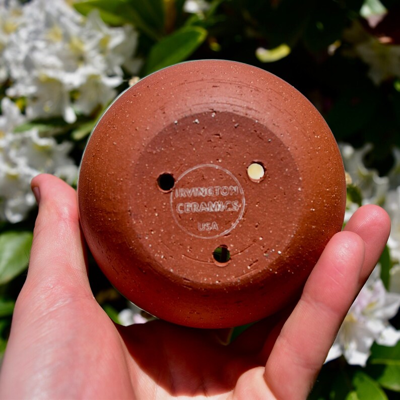 Puede incluir: Una maceta de cer&aacute;mica de color terracota con tres agujeros de drenaje. La parte inferior de la maceta tiene una etiqueta blanca que dice "IRVINGTON CERAMICS USA".