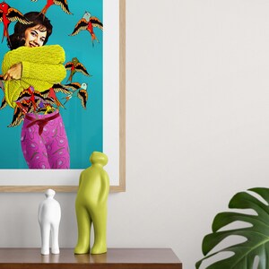 May include: A framed print of a woman wearing a yellow sweater and pink pants with a blue background and many colorful birds flying around her. Two abstract sculptures are on a wooden table in front of the print. A large green leaf plant is on the right side of the image.