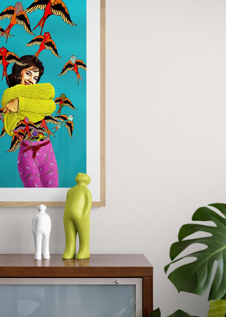 May include: A framed print of a woman wearing a yellow sweater and pink pants with a blue background and many colorful birds flying around her. Two abstract sculptures are on a wooden table in front of the print. A large green leaf plant is on the right side of the image.