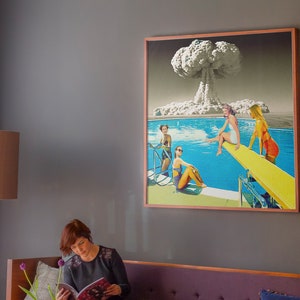 May include: A framed collage artwork depicting a mushroom cloud explosion above a swimming pool with four women in swimsuits. The women are on a diving board and in the pool. The artwork is in a wooden frame.