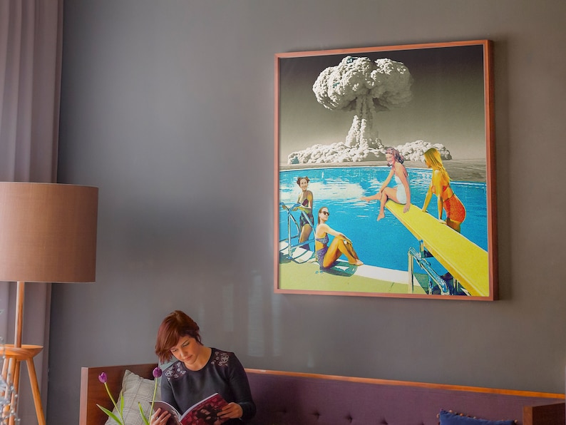 May include: A framed collage artwork depicting a mushroom cloud explosion above a swimming pool with four women in swimsuits. The women are on a diving board and in the pool. The artwork is in a wooden frame.