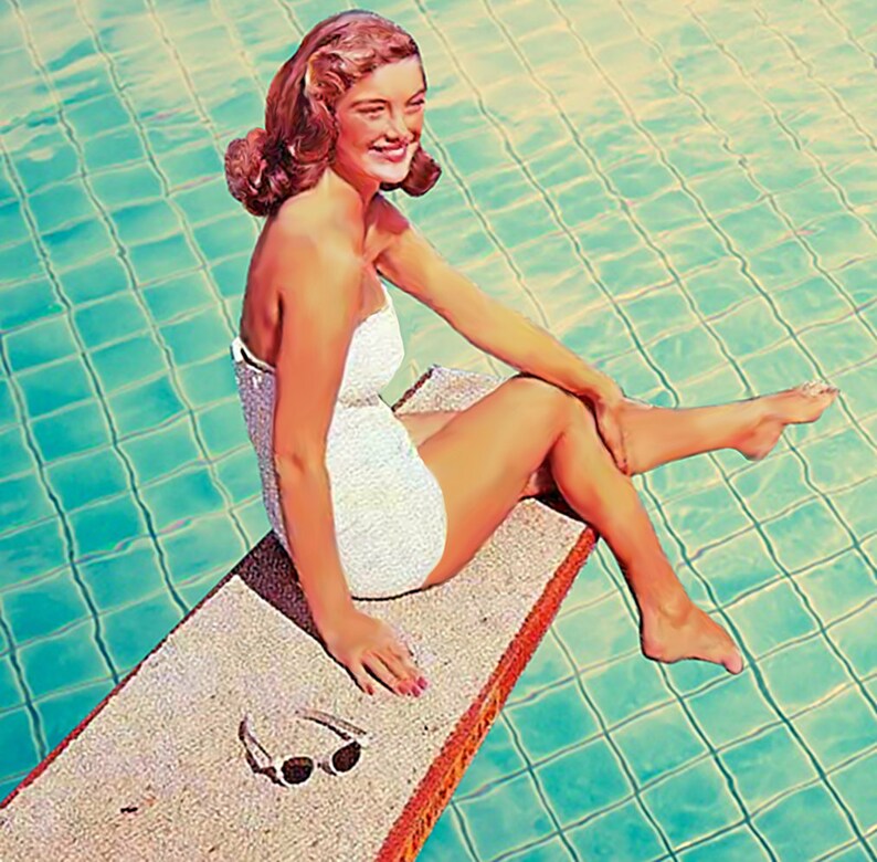 May include: A woman in a white swimsuit sits on a diving board by a swimming pool. The pool water is a bright turquoise color.