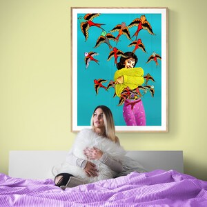 May include: A woman sits on a bed with a purple and white striped comforter, holding a white fluffy pillow. Behind her is a framed print of a woman in a yellow sweater and pink pants surrounded by colorful birds on a turquoise background.