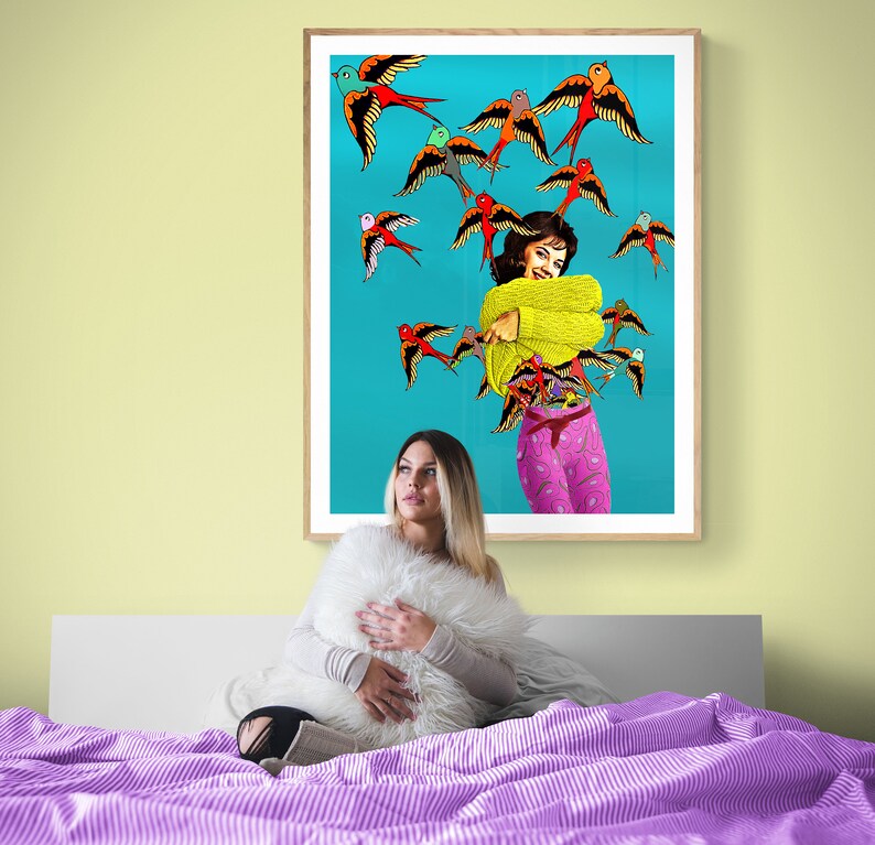 May include: A woman sits on a bed with a purple and white striped comforter, holding a white fluffy pillow. Behind her is a framed print of a woman in a yellow sweater and pink pants surrounded by colorful birds on a turquoise background.