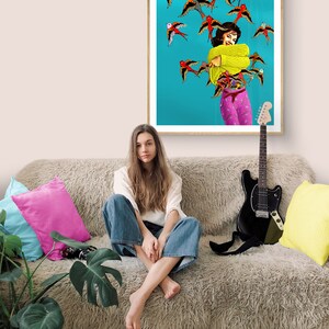 May include: A woman sits on a fluffy couch with a guitar and a framed print of a woman surrounded by colorful birds. The woman in the print is wearing a yellow sweater and pink pants.