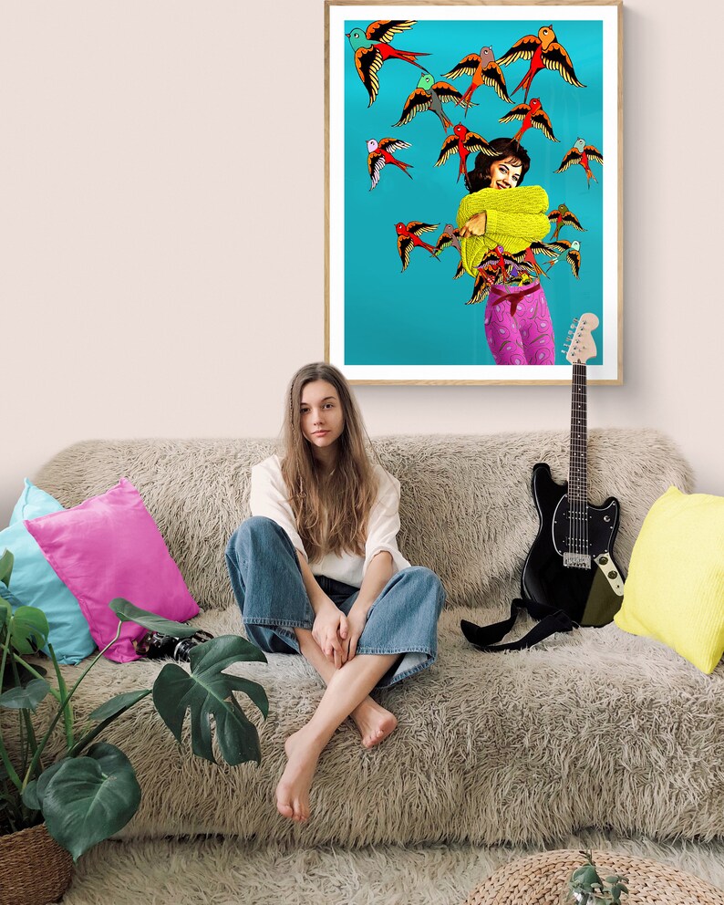 May include: A woman sits on a fluffy couch with a guitar and a framed print of a woman surrounded by colorful birds. The woman in the print is wearing a yellow sweater and pink pants.