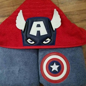 Puede incluir: Una toalla con capucha roja con un emblema de Capit&aacute;n Am&eacute;rica azul y blanco y alas blancas. La toalla est&aacute; doblada sobre una toalla azul con un emblema de escudo del Capit&aacute;n Am&eacute;rica.