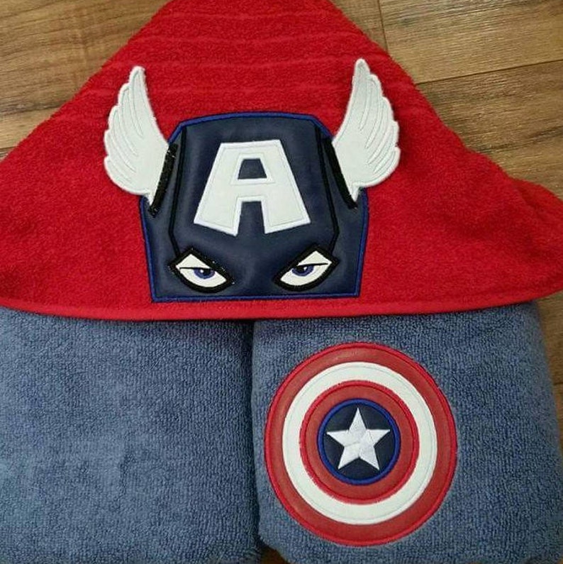 Puede incluir: Una toalla con capucha roja con un emblema de Capit&aacute;n Am&eacute;rica azul y blanco y alas blancas. La toalla est&aacute; doblada sobre una toalla azul con un emblema de escudo del Capit&aacute;n Am&eacute;rica.