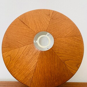 Puede incluir: Una pantalla de l&aacute;mpara redonda de madera con un acabado marr&oacute;n claro. La pantalla tiene una abertura central para una bombilla y un anillo de metal blanco alrededor de la abertura.