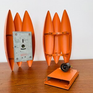 May include: Two orange metal wall sconces with a unique design. The sconces are made of five curved metal pieces that are joined together. The sconces are shown disassembled with a metal back plate and a black electrical socket.