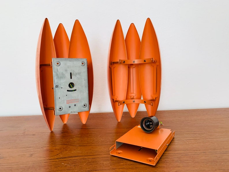 May include: Two orange metal wall sconces with a unique design. The sconces are made of five curved metal pieces that are joined together. The sconces are shown disassembled with a metal back plate and a black electrical socket.