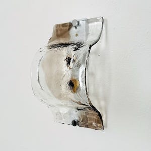 May include: A clear and brown glass wall sconce with a unique, organic shape. The sconce is mounted on a white wall with two silver metal brackets. The glass has a textured appearance and a dark brown tint on the edges.