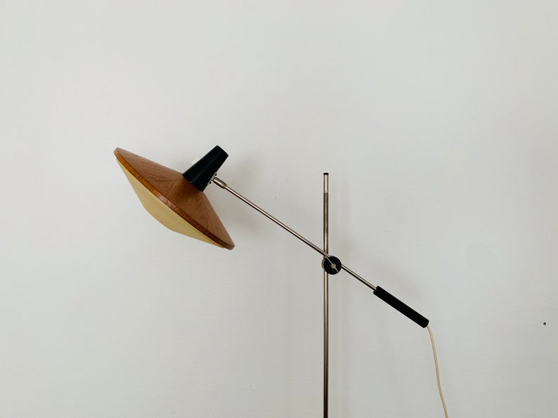 Puede incluir: Una l&aacute;mpara de pie moderna de mediados de siglo con una pantalla de madera y un brazo de metal plateado. La l&aacute;mpara tiene una base negra y un cable blanco.