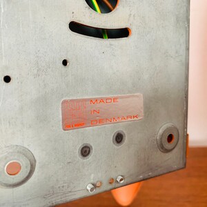 May include: Close-up of a metal back panel with a sticker that reads "MADE IN DENMARK". The panel has several holes and is a light gray color.