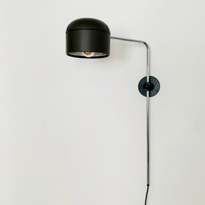 May include: A black wall-mounted lamp with a round shade and a long, silver arm. The lamp is attached to the wall with a black circular base.