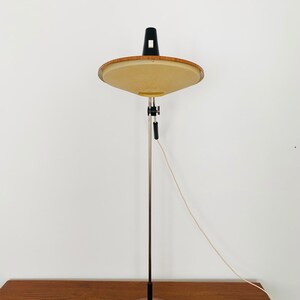 Puede incluir: Una l&aacute;mpara de pie moderna de mediados de siglo con una base de madera y una pantalla redonda de color beige. La l&aacute;mpara tiene un brazo de metal negro y un cable blanco.