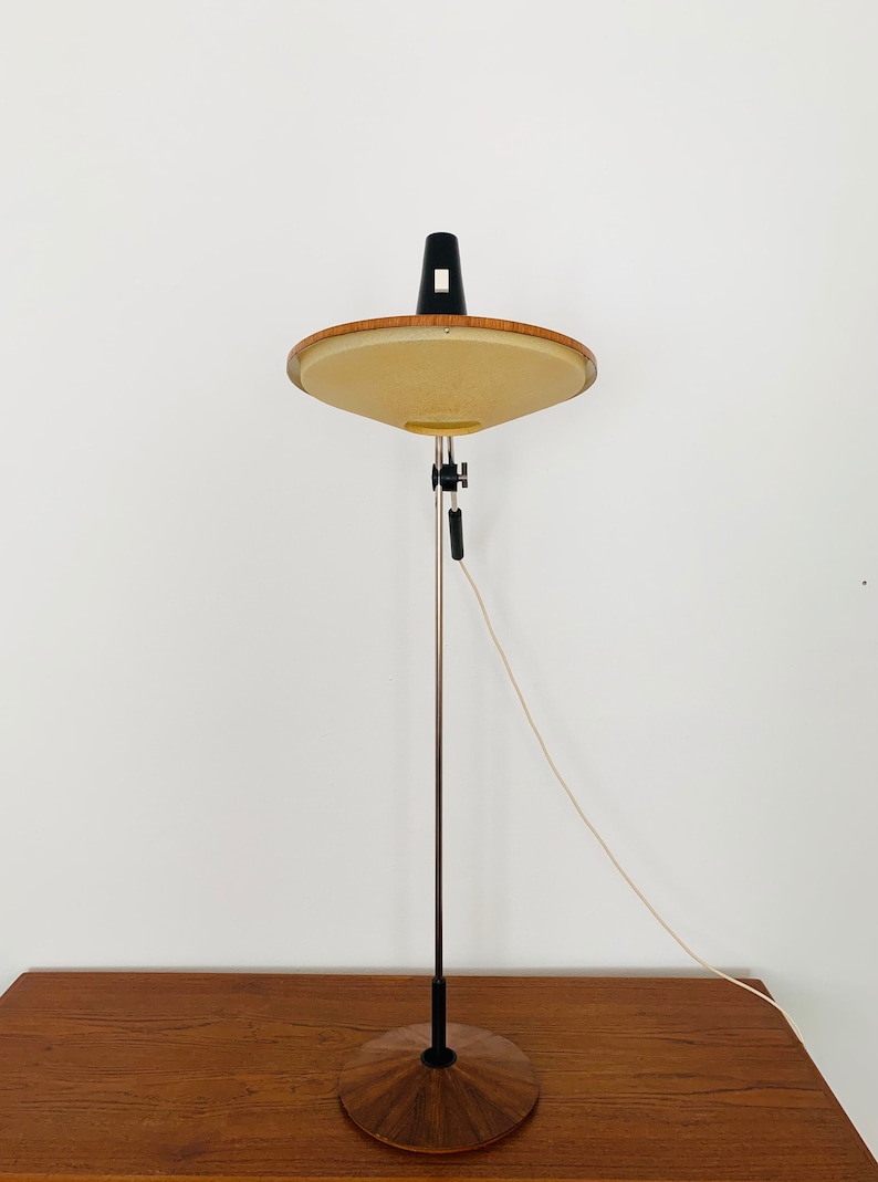 Puede incluir: Una l&aacute;mpara de pie moderna de mediados de siglo con una base de madera y una pantalla redonda de color beige. La l&aacute;mpara tiene un brazo de metal negro y un cable blanco.