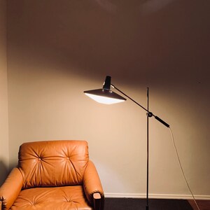 Puede incluir: Un sill&oacute;n de cuero marr&oacute;n se encuentra frente a una l&aacute;mpara de pie negra con una pantalla blanca y una base de madera. La l&aacute;mpara est&aacute; encendida y proyecta luz sobre el sill&oacute;n.