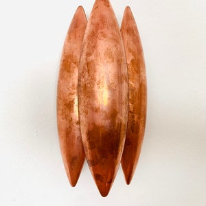 May include: Three copper wall sconces in the shape of elongated teardrops, mounted on a white wall.