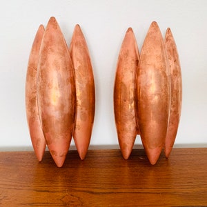 May include: A pair of copper wall sconces with a unique design featuring multiple curved, elongated shapes. The sconces are made of a shiny, metallic copper and have a modern, minimalist aesthetic.