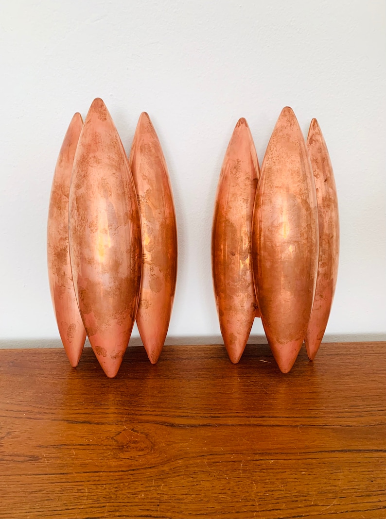 May include: A pair of copper wall sconces with a unique design featuring multiple curved, elongated shapes. The sconces are made of a shiny, metallic copper and have a modern, minimalist aesthetic.