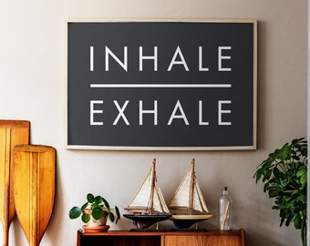 Könnte beinhalten: Ein schwarzer gerahmter Druck mit weißem Text, der "INHALE EXHALE" lautet, hängt an einer beigefarbenen Wand über einem Holzregal mit zwei dekorativen Segelbooten, einer Topfpflanze und einer Glasflasche.