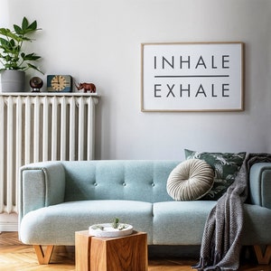 Op de afbeelding: Een lichtblauwe bank met een grijze deken en een rond grijs kussen. De bank staat voor een witte radiator en een ingelijst print met de tekst "INHALE EXHALE".